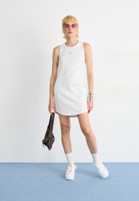 Hvit ribbestrikket ermeløs kjole med rund hals og en liten svart logo. Modellen holder en mørk veske, iført platå sneakers og hvite sokker.