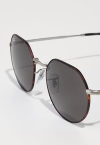 Ochelari de soare Ray-Ban cu un cadru hexagonal din acetat cu model țestoasă maro, lentile închise, accente metalice argintii și tampoane pentru nas transparente.