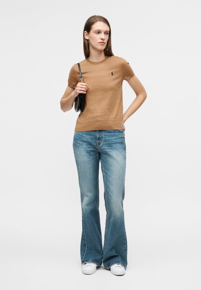 Καφέ κοντομάνικη πλεκτή μπλούζα με ριμπ λαιμόκοψη και μικρό λογότυπο, σε συνδυασμό με φαρδιά ανοιχτό μπλε τζιν παντελόνια και λευκά αθλητικά παπούτσια.