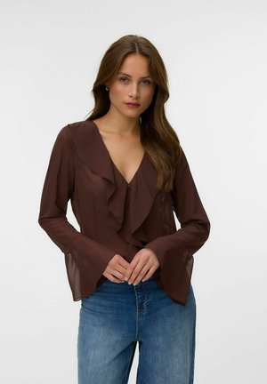Kvinde med langt brunt hår iført en gennemsigtig brun flæsebluse og blå jeans, stående mod en ensfarvet hvid baggrund.