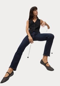 Chaleco negro sin mangas con botones, jeans azul oscuro y zapatos planos negros con lunares blancos. Modelo sentado con las piernas estiradas y un fondo neutro.
