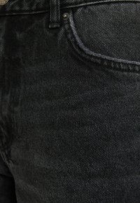 Gros plan sur un jean en denim noir montrant la poche avant, les coutures, la boucle de ceinture et les détails des rivets en métal.