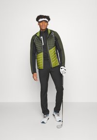 Sporty golfoutfit bestående af en tofarvet grøn og sort isoleret jakke, sorte tætsiddende bukser og hvid/blå golfsko. Holder et golfjern.