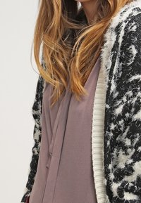 Schwarzer und weißer gemusterter Cardigan mit flauschiger Textur und cremefarbenem Besatz, getragen über einem hellen taupefarbenen Top mit V-Ausschnitt und Faltendetail.