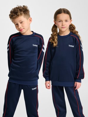 Zwei Kinder in dunkelblauer Sportkleidung mit roten Paspeln und weißem Logo, die nebeneinander vor einem einfarbigen Hintergrund stehen.