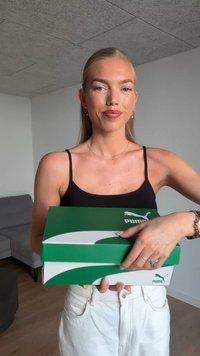 Zielone pudełko po butach Puma z białym logo i wzorem, trzymane przez osobę ubraną w czarną górę i białe spodnie, na jasnym tle.