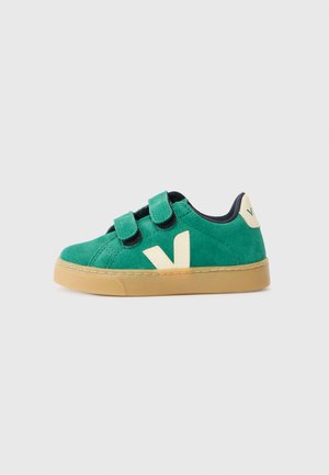 Grønn semsket sneakers med borrelås, beige detaljer, gummisåle og en hevet bokstav "V"-logo på siden. Utsmykket med en marineblå innside.