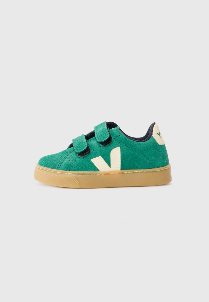 Sapatilha verde de camurça com correias de velcro, detalhes em bege, sola de borracha e um logótipo em relevo da letra "V" na lateral. Com interior azul-marinho.