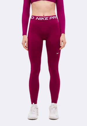 Le modèle porte un legging Nike Pro magenta et un crop top assorti à manches longues, ainsi que des baskets blanches. Il se tient devant un fond uni.