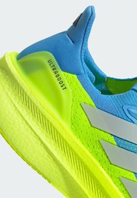 Zapatilla deportiva azul y verde neón con un upper texturizado, tres franjas plateadas y una suela acolchada marcada como "ULTRABOOST."