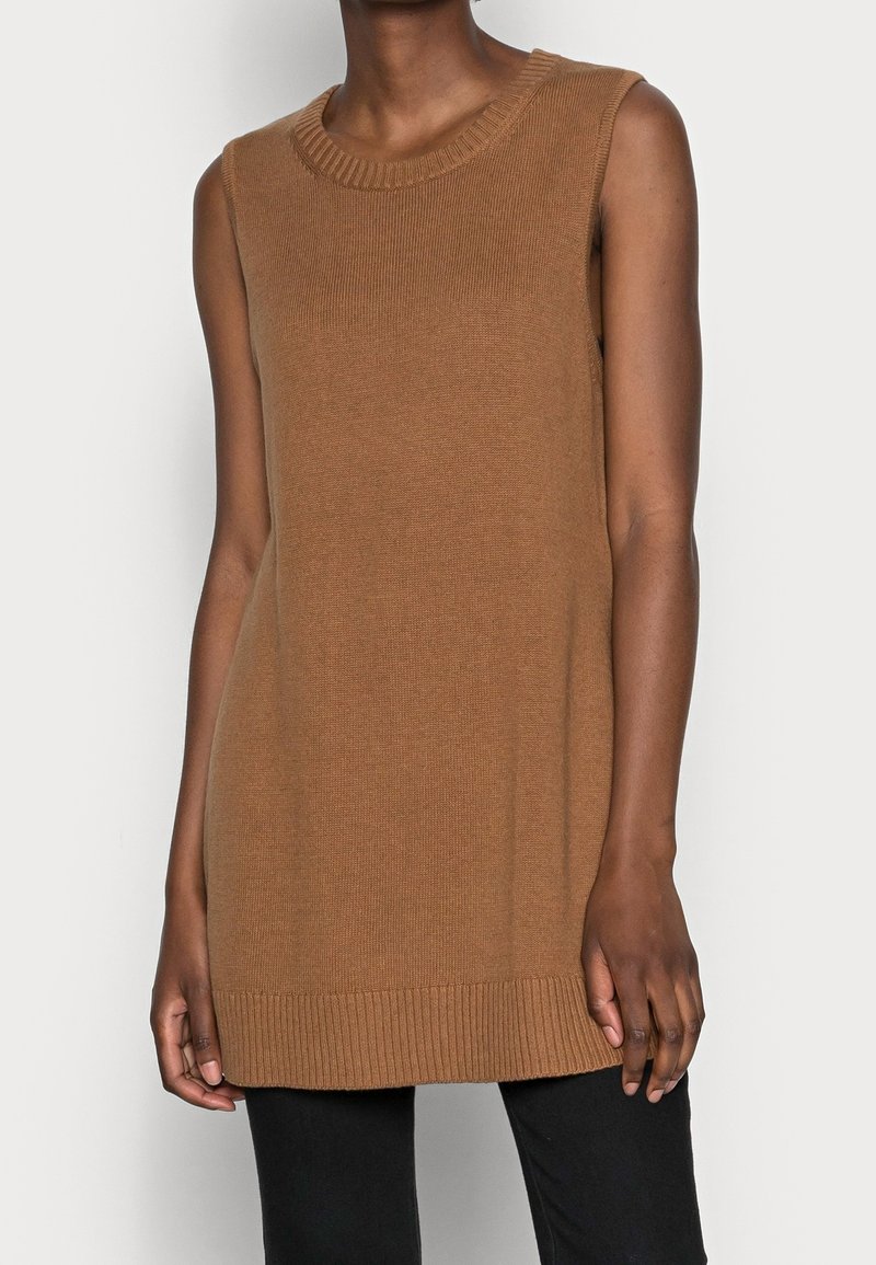 Veste sans manches en tricot marron portée sur un pantalon noir, visible des épaules jusqu'au milieu des cuisses sur une personne debout.