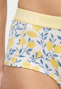 Κίτρινα σλιπ με μπλε φύλλα και λεμονόμορφο μοτίβο, φορεμένα στον γοφό, με το όνομα της μάρκας "PIECES" στη μέση.