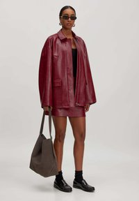 Burgunderrote Lederjacke mit übergroßem Design und zwei Fronttaschen, kombiniert mit einem passenden Minirock und einer grauen Wildleder-Schultertasche. Schwarze Schuhe und Sonnenbrille.