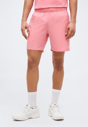 Mann trägt kniehohe rosa Sportshorts, weiße Kniestrümpfe und weiße Sportschuhe und steht vor einem schlichten Hintergrund.