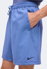 Shorts de sport bleus avec une taille élastique et un cordon, portés par une personne avec une main dans la poche et un logo Nike noir près de l'ourlet.