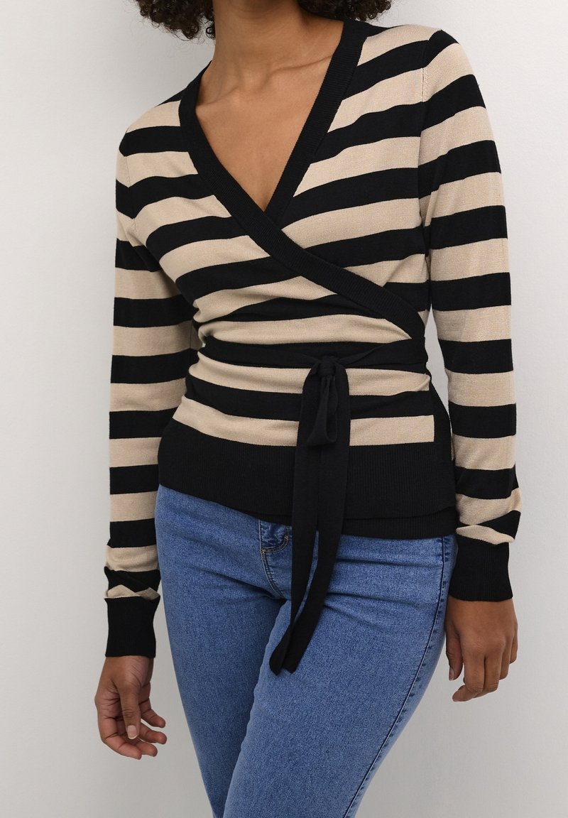 Femme portant un pull cache-cœur à rayures horizontales beige et noires, noué à la taille, associé à un jean bleu sur un fond uni.