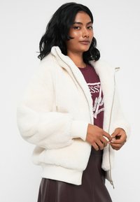 Donna con capelli neri che indossa una giacca zip bianca e soffice sopra una maglietta bordeaux e una gonna scura, su uno sfondo bianco.