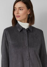 Graue Wildlederjacke mit einem Kragen, vorderen Knöpfen und langen Ärmeln.getragen über einem weißen gerippten Rollkragenpullover mit einer glatten Textur.