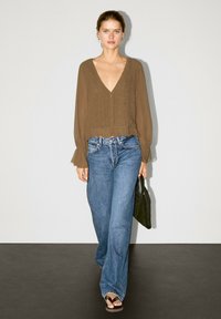 Braune, strukturierte Bluse mit V-Ausschnitt, kombiniert mit lockeren blauen Jeans und schwarzen Sandalen. Das Modell hält eine dunkelgrüne Tasche. Neutraler Hintergrund.