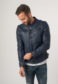 Giubbotto in pelle navy da motociclista con colletto rialzato, tasche con zip e maniche attillate, abbinato a jeans chiari scoloriti.