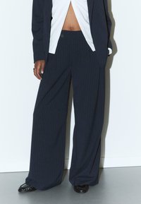 Marineblau- und rot gestreifte weite Hosen mit hoher Taille, die über einen einzelnen Knopfverschluss und eine glatte Textur verfügt. Kombiniert mit einem weißen Hemd.
