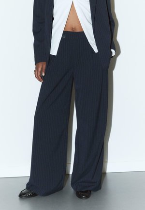 Marineblau- und rot gestreifte weite Hosen mit hoher Taille, die über einen einzelnen Knopfverschluss und eine glatte Textur verfügt. Kombiniert mit einem weißen Hemd.