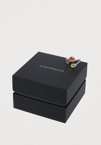 Ασημένιο δαχτυλίδι με τρεις πολύχρωμους λίθους – μωβ, πορτοκαλί και κίτρινο – τοποθετημένο σε μαύρο κουτί με λογότυπο, που παρουσιάζει ένα κομψό, μοντέρνο σχέδιο.