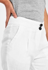 Pantalons en lin blancs à taille haute, dotés de deux boutons noirs et de poches latérales. Texture lisse avec un éclat subtil.