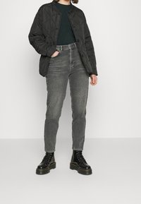 Personne portant une veste matelassée noire, un pull vert foncé, un jean gris taille haute et des bottines à lacets noires sur plateforme, se tenant avec une main dans la poche.