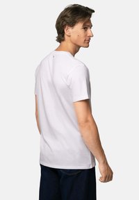 Ung mand med kort brunt hår iført en ensfarvet hvid kortærmet T-shirt og mørkeblå bukser, vist fra ryggen i en neutral omgivelser.