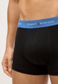 Boxer briefs de algodón negro con una cinturilla elástica azul que presenta el logotipo y emblema de "TOMMY HILFIGER". Textura suave, diseño ajustado.