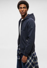 Tamnoplava zip-up hoodie od mekog materijala, s kapuljačom na vezanje, bočnim džepovima i zakrpu na rukavu. Uparena s kariranim košuljom vezanom oko struka.