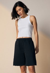 Weißes, kurzes Tanktop aus Baumwolle, kombiniert mit schwarzen Shorts mit elastischem Bund und ausgefranstem Saum. Glatte Textur, lässige Passform.