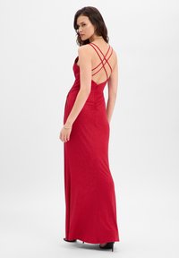 Rode jurk tot op de grond met kruisbanden aan de achterkant, geplooide zijdetails en een glinsterende textuur, gecombineerd met schoenen met hoge hakken.