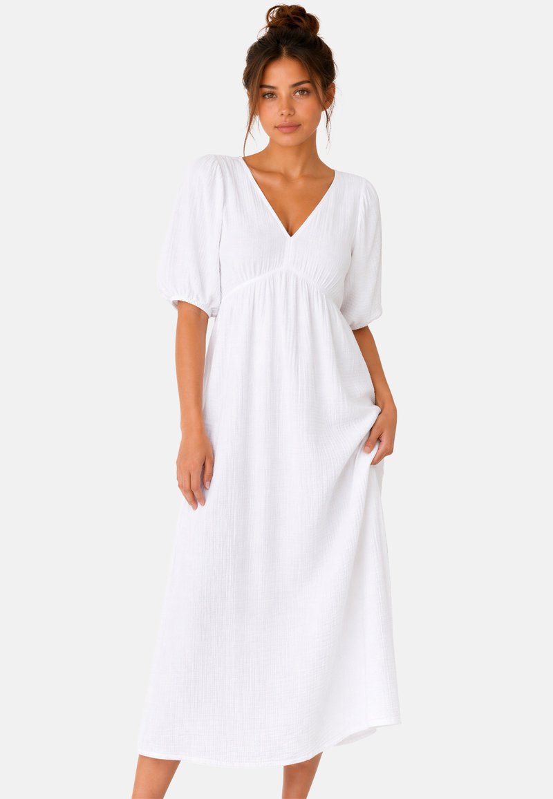 Donna con capelli castani raccolti in uno chignon che indossa un vestito midi bianco con scollo a V e maniche corte gonfie, in piedi davanti a uno sfondo semplice.