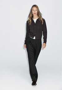 Cardigan marron foncé avec une fermeture à boutons, associé à une chemise blanche et un pantalon noir à rayures. Les chaussures présentent un design à lanières.