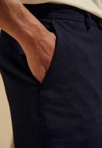 Pantalon texturé bleu marine avec une coupe ajustée, comprenant une poche latérale. Main visible reposant dans la poche, mettant en valeur le tissage du tissu.