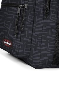 Sac à dos noir avec un tissu texturé comportant des motifs géométriques sombres, des compartiments zippés et une étiquette de marque. Poche latérale en mesh visible.