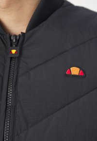 Gros plan sur une personne portant une veste matelassée noire avec une fermeture éclair partiellement ouverte à l'avant et un petit logo orange et rouge sur la poitrine.