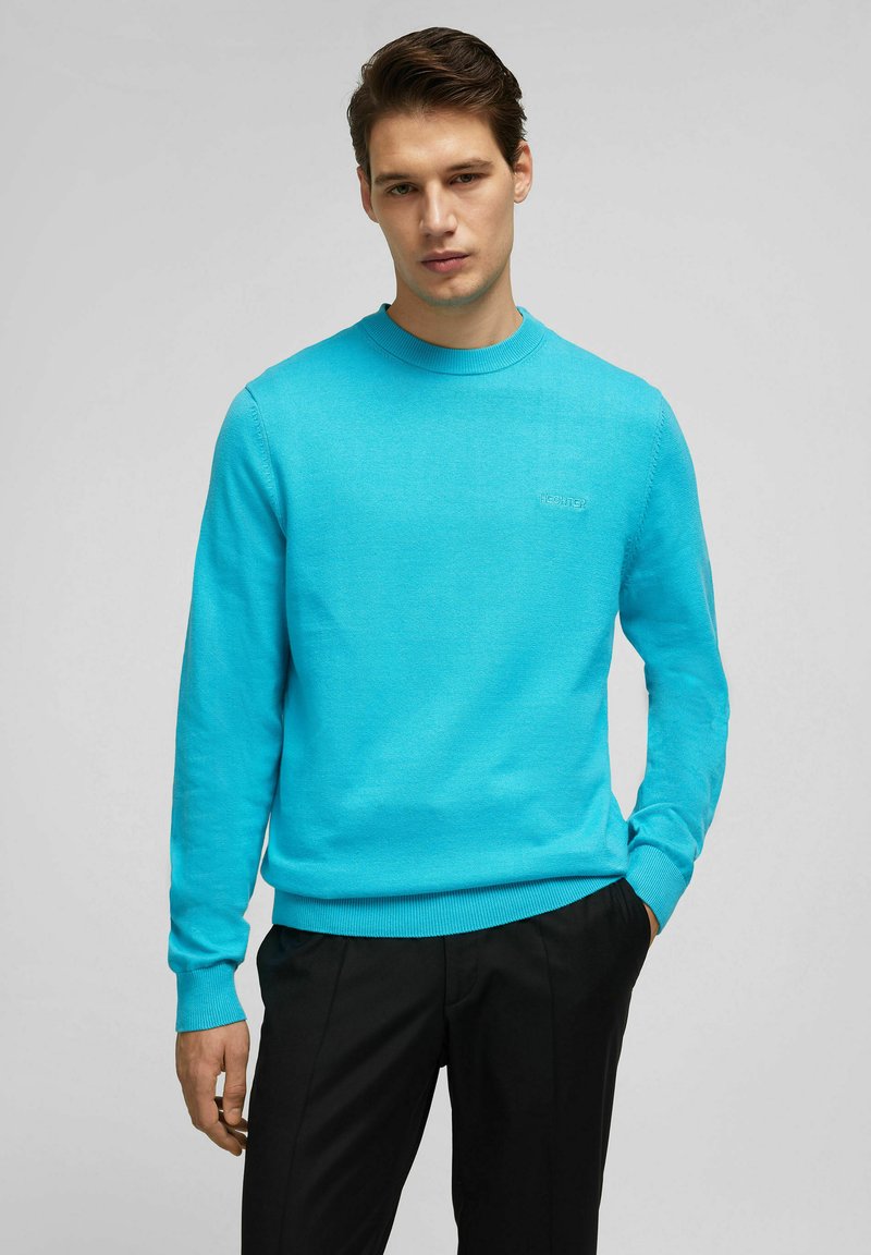 Türkiser Pullover mit Rundhalsausschnitt, gerippten Bündchen und Saum. Mit einem dezenten Logo auf der Brust. Kombiniert mit schwarzen Hosen.