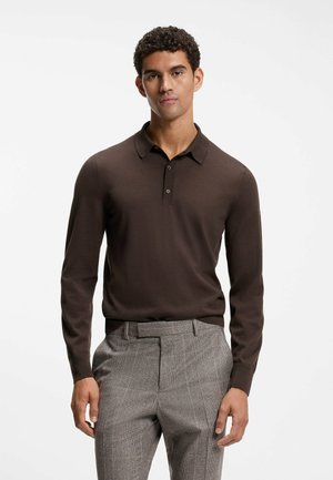 Mand iført en mørkebrunt langærmet poloshirt, stoppet ned i grå skræddersyede bukser, stående mod en ensfarvet hvid baggrund.
