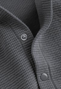 Close-up of grey waffle-knit fabric with two metal snap buttons, one open and one closed, showing texture and fastening details.