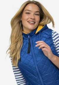 Modrý prošívaný vest s žlutou kapucí a černým zipem. Pod ním je vidět pruhované triko s dlouhým rukávem. Hladká textura a přiléhavý střih.