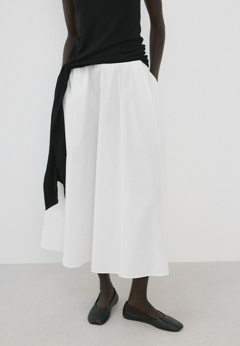 Žena v černém topu bez rukávů a splývavé bílé midi sukni s kapsami, doplněná černými balerínami, stojící před jednobarevným pozadím.