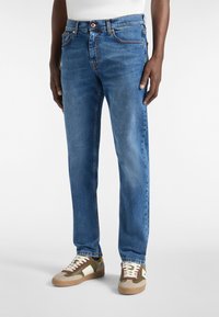 Vaqueros de mezclilla azul con un corte recto, cinco bolsillos, costuras naranjas y una cintura con botón. Combinados con zapatillas beiges y aceitunas.