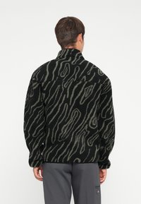 Black and olive green patterned fleece jacket with a high collar and oversized fit, featuring wavy lines and a textured surface.