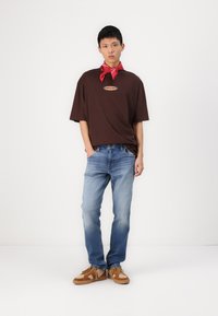 Camiseta marrón de gran tamaño con un logo, jeans azul, y zapatillas beige con acentos texturizados. Pañuelo rojo atado alrededor del cuello.
