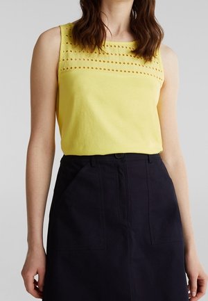 Frau trägt ärmelloses gelbes Oberteil mit perforierten horizontalen Streifen über der Brust und einen hoch taillierten schwarzen Rock mit Taschen.