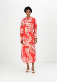 Rochie roșie cu mâneci lungi, cu un model floral alb, butoane pe toată lungimea din față și o siluetă fluidă. Stilizată cu sandale neutre, ușor de purtat.
