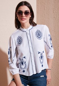 Chemisier blanc à boutons avec broderie florale bleu marine, manches 3/4 et col rond, associé à un short en denim.